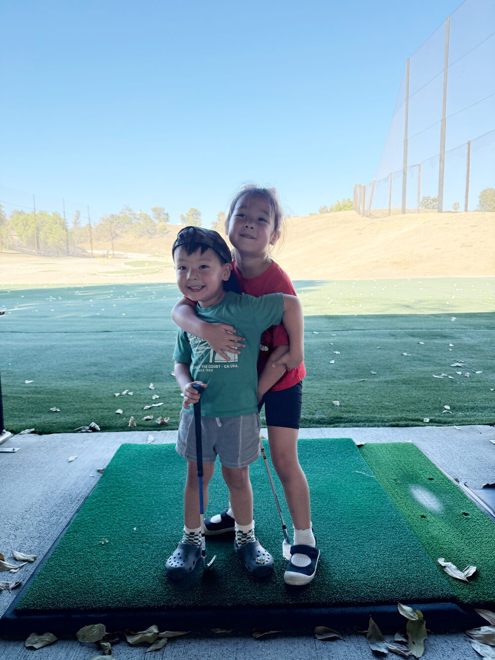 Two young siblings share a hug on the driving range mat after a kids golf lesson, clubs in hand