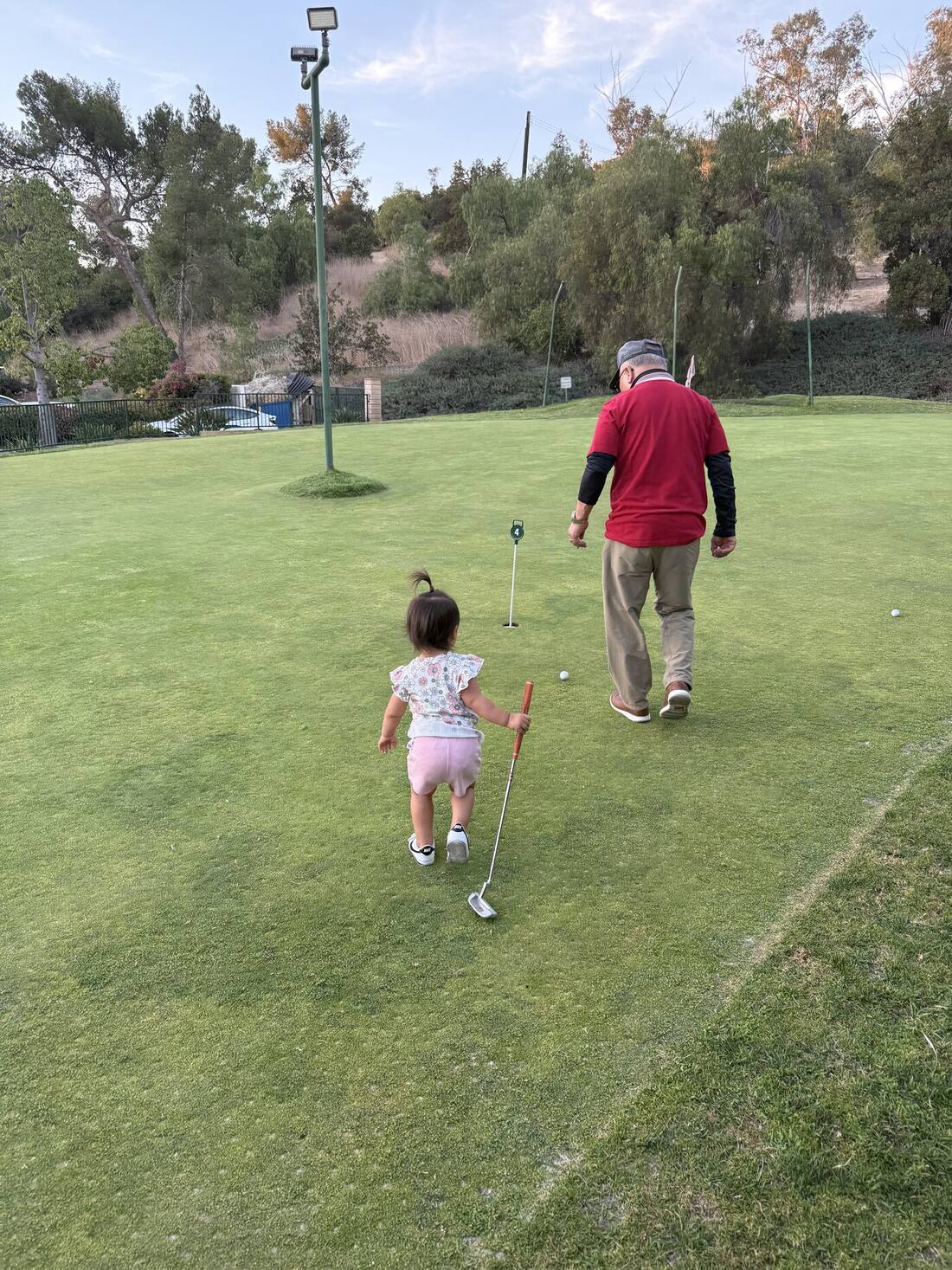 A toddler and her grandfather walking the real-grass putting green at dusk
