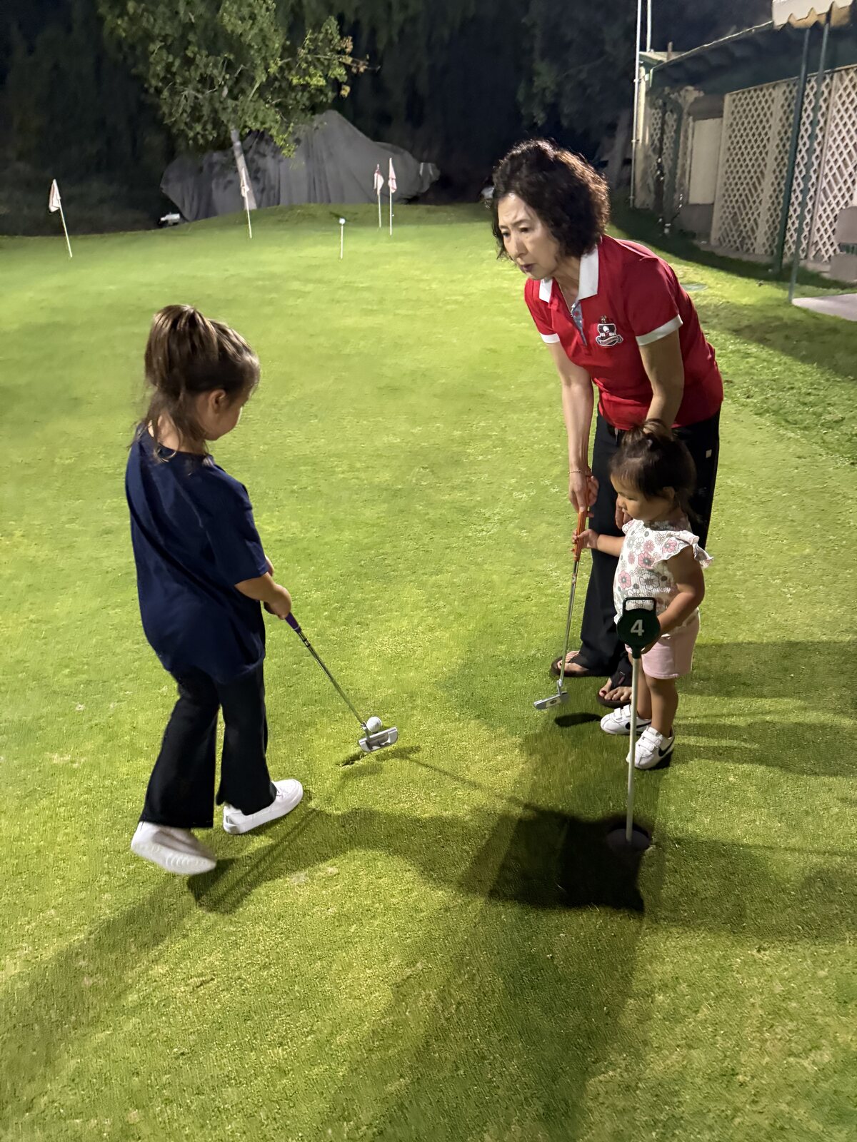 A grandmother helps two young girls with their putting on the real-grass course at night